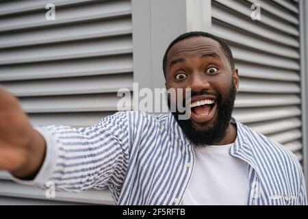 Close up portrait of excited African American man taking selfie by mobile phone standing on urban street. Emotional blogger influencer shooting video Stock Photo