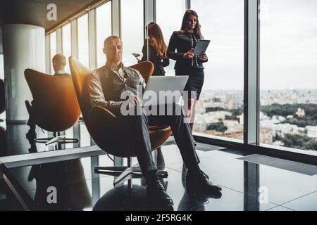 A bossy caucasian man entrepreneur on an orange armchair is using his laptop while a businesswoman with a tablet pc in hands is standing aloof next to Stock Photo