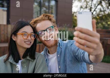 Happy smiling friends influencers using mobile phone, recording video outdoors. woman and red man wearing stylish eyeglasses taking selfie Stock Photo