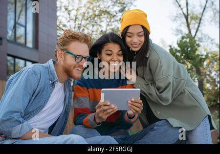 Group of smiling multiethnic friends using digital tablet, watching video, shopping online, sitting outdoors. University students studying online Stock Photo