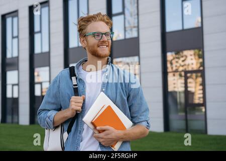Smiling university student with backpack and book walking in university campus, education concept. Portrait of handsome man wearing stylish eyeglasses Stock Photo