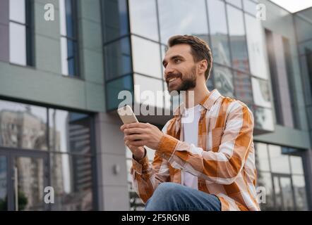 Young man using mobile phone, working online, sitting outdoors. Successful freelancer holding smartphone, receive payment Stock Photo