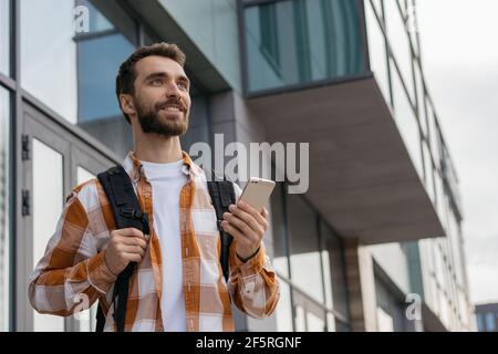 Smiling handsome man using mobile phone outdoors. Young bearded tourist with backpack walking on urban street, searching best way. Travel concept Stock Photo