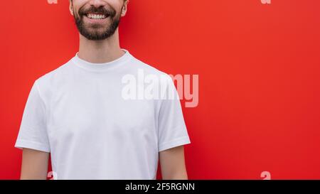 Young handsome model wearing casual t-shirt isolated on red background, mock up. Cropped image of stylish bearded man smiling Stock Photo