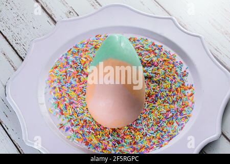 White chocolate Easter egg painted in green, orange and pink. On an old plate covered with colorful chocolate sprinkles. Stock Photo