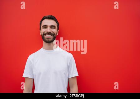 Portrait of bearded man with happy emotional face looking at camera, smiling. Young handsome model wearing casual t-shirt isolated on red background Stock Photo