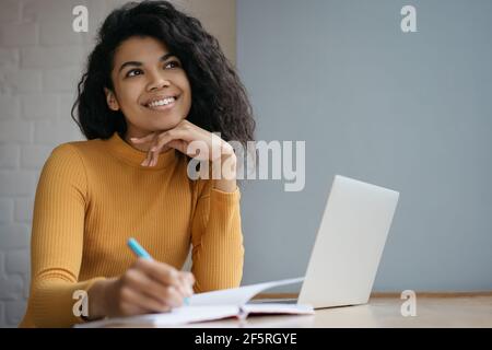 Portrait of happy pensive student studying, exam preparation, online education. Beautiful African American woman  taking notes working from home Stock Photo