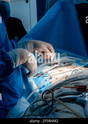 A doctor in a blue uniform uses medical instruments and metal thread to suture the skin during minimally invasive surgery. Close up hands in sterile Stock Photo