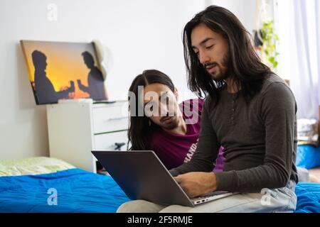 twin brothers lying in bed looking at the computer, 2 long-haired men looking at the computer, lying on the bed Stock Photo