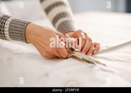 Close up on hands of unknown caucasian woman holding scissors cutting textile on the table at home or studio - hobby and tailoring concept Stock Photo