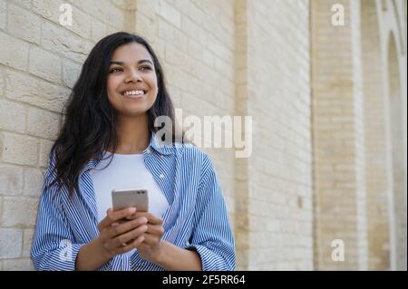 Young smiling woman using mobile phone shopping online looking away, copy space. African American freelancer holding smartphone, receive payment Stock Photo