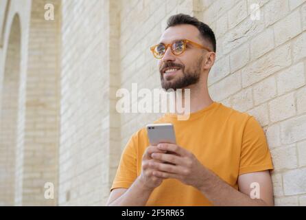 Young handsome man using mobile phone shopping online looking away standing on the street. Confident freelancer in stylish eyeglasses receive payment Stock Photo