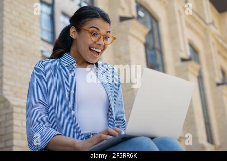 Excited African American woman using laptop computer shopping online with sales. Happy beautiful female watching video, reading good news Stock Photo