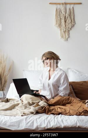 Woman Concentrated Looks At Computer Screen. Freelancer In White Blouse And glasses Using Laptop. Work From Home Freelance Lifestyle Concept. Stock Photo