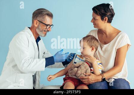 Doctor giving flu shot of a small boy sitting with his mother in clinic. Boy getting vaccinated by a pediatrician. Stock Photo