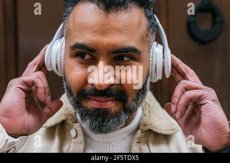 Crop friendly bearded ethnic male listening to song from wireless headset while looking away in daylight Stock Photo