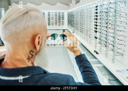 Back view elderly bearded male in trendy outfit standing with modern stylish sunglasses in optical shop Stock Photo