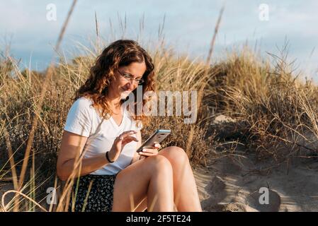 Content female in summer outfit sitting on sandy shore and messaging on smartphone while enjoying sunset Stock Photo