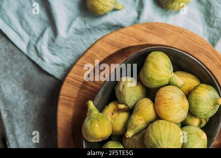 Fresh and ripe sweet green figs, in a black bowl on the wood plate served on the blue tablecloth table, Seasonal organic fruit. Also known as ripe whi Stock Photo