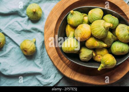 Fresh and ripe sweet green figs, in a black bowl on the wood plate served on the blue tablecloth table, Seasonal organic fruit. Also known as ripe whi Stock Photo