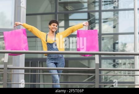 Delighted African American female standing with paper bags in city and looking at camera after successful shopping Stock Photo