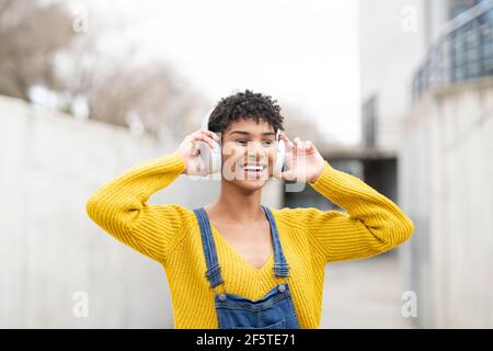 Dreamy smiling African American female in wireless headphones enjoying songs while standing on street Stock Photo
