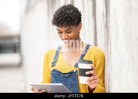 Glad black female standing on street with coffee to go in paper cup and watching video on tablet while entertaining at weekend in city Stock Photo