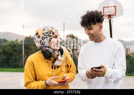 Delighted diverse friends standing on basketball playground and browsing smartphones while laughing and having fun in city Stock Photo
