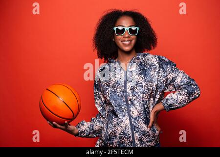 Trendy African American female in street style wear and cool sunglasses standing with basketball ball in hands against red background Stock Photo