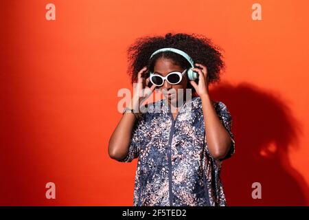 Cheerful young African American female in stylish outfit listening to cool music in wireless headphones and looking away with sunglasses against red w Stock Photo