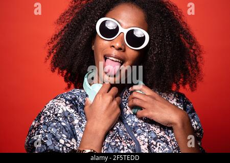 Cool young African American female in trendy outfit and sunglasses carrying wireless headphones around neck and showing tongue against red background Stock Photo