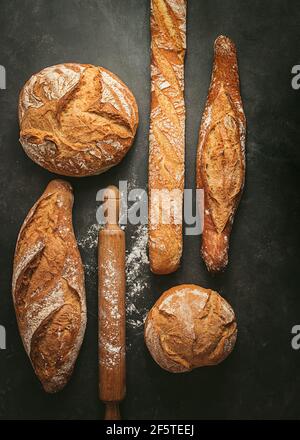 Top view composition with various typed of freshly baked crusty artisan bread loaves of different shapes placed near wooden rolling pin on black backg Stock Photo