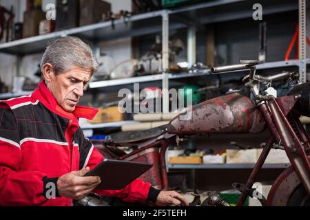 Serious gray haired elderly repairman using tablet and watching video instruction while fixing old broken motorcycle in garage Stock Photo