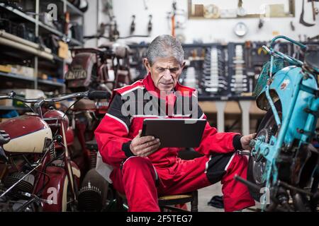 Serious gray haired elderly repairman using tablet and watching video instruction while fixing old broken motorcycle in garage Stock Photo