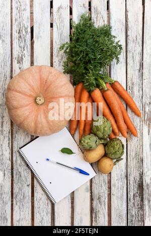 Top view of harvest of assorted fresh vegetables and opened notepad with pen arranged on wooden table Stock Photo