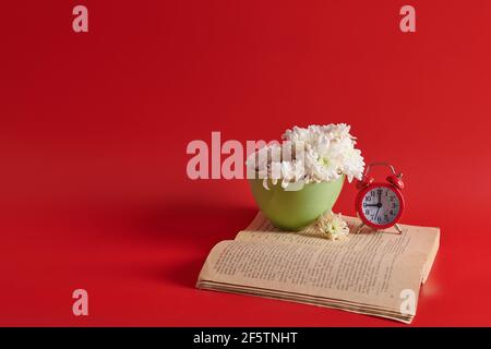 stand on a book white flowers in a cup with an alarm clock on a red background Stock Photo