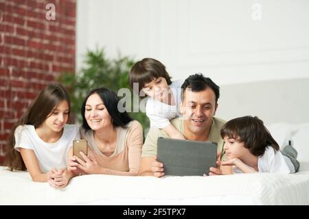 Boys and girls. Happy latin family with cute kids using digital devices, lying on the bed together Stock Photo