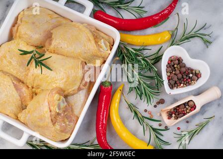 Raw marinated chicken thigh in yellow marinade in a white oven baking dish with fresh rosemary sprigs and assorted spices on a marble kitchen board. T Stock Photo