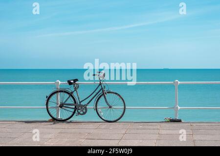 Grey bicycle leaning against white railings with pink paving in foreground and turquoise sea and blue sky behind. Stock Photo