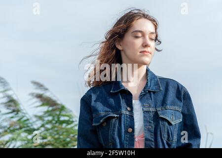 Teenage girl outside eyes closed in tranquil state, wearing denim jacket face turned to the sun. Soft focus tall grass in background. Stock Photo