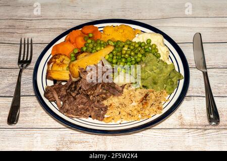 Sunday dinner of slow cooked shredded pulled beef on a plate with a knife and fork and plenty of vegetables such as peas, mushy peas, parsnips, roast Stock Photo