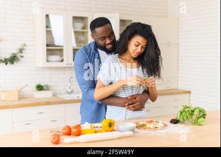 Loving black husband hugs wife who preparing his favorite food - homemade delicious pizza, hungry man waiting for a tasty yummy lunch, watches his wife decorates the dough with greenery and filling Stock Photo