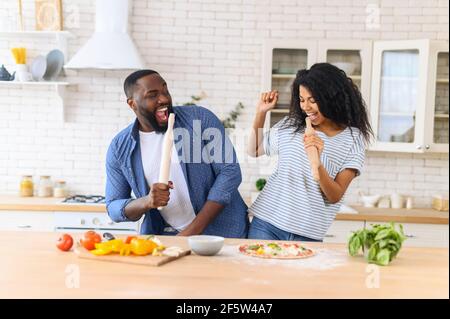 Happy multiethnic family in the kitchen having fun and cooking together, healthy food at home, young joyful couple have fun dancing and singing with rolling pin while set the table for breakfast Stock Photo