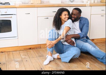 Married couple sitting on warm wooden floor in modern kitchen, hugging looking at each other, family enjoying weekend at home together, posing for photo together, holiday concept during the weekend Stock Photo