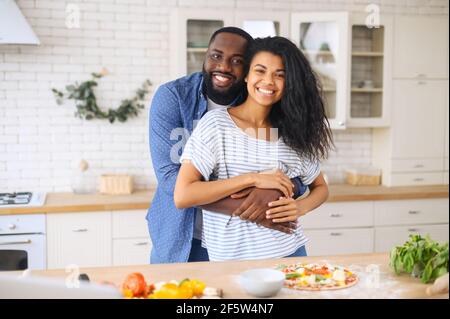 Multi ethnic family with a loving husband hugging his wife from behind in the kitchen cooking pizza together, right preparation, make yummy home-made pizza, enjoy communication, cookery hobby concept Stock Photo