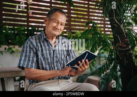 Happy senior thai man sitting on marble chair under the tree and reading book, Healthy senior concept Stock Photo