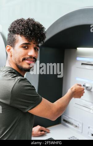 latin american young man looking at camera and smiling while using an ATM Stock Photo