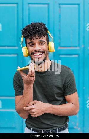 portrait of an attractive young man smiling and looking at camera. He is holding a yellow smart phone and wears a yellow headphones. He is standing on the street and with a blue background Stock Photo