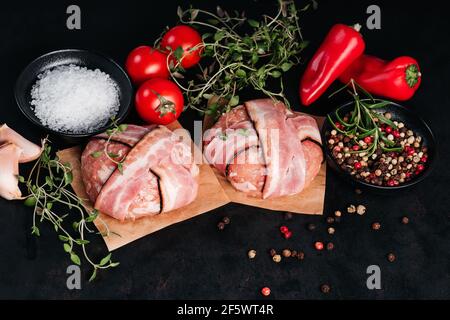 Two chicken hamburgers wrapped in bacon on cave paper with rosemary branches, black pepper, sweet red peppers, tomatoes and garlic on a black backgrou Stock Photo