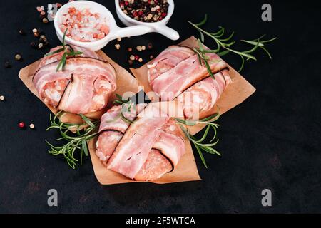 Three chicken hamburgers wrapped in bacon on cave paper with rosemary branches, black pepper and pink salt in white small plates, black background. Fa Stock Photo